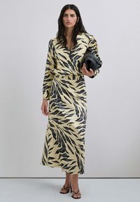 Vestido largo y ajustado en amarillo con rayas de tigre negras. Presenta un escote en V y mangas largas. Combinado con un bolso de mano negro y sandalias con tiras.