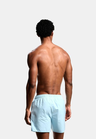 Άνδρας με σκουρόχρωμο δέρμα, κοντά κατσαρά μαλλιά, γυμνός από τη μέση και πάνω, με ανοιχτό μπλε μαγιό, γυρισμένος πλάτη, σε απλό ανοιχτό φωτεινό φόντο.