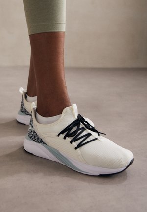 Personne portant des chaussures de sport blanches avec des lacets noirs et un talon à motif, debout sur un sol gris lisse.