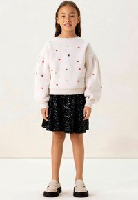 Lichtgrijze sweatshirt met kleurrijke borduurwerk van sterren, harten en een maan, gecombineerd met een zwarte paillettenrok en beige instapschoenen met dikke zolen.