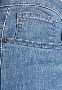 Tela de mezclilla azul que presenta un bolsillo trasero con detalles de costura y un remache de metal. La textura parece ligeramente desgastada y descolorida.