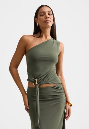 Žena v olivovo zelenej asymetrickej blúzke s uzlom a zladenou nariasenou sukňou, doplnená zlatými náušnicami a jantárovým náramkom.