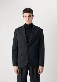 Giacca nera con rever a punta, un solo bottone e due tasche laterali. Indossata sopra un dolcevita nero su uno sfondo chiaro e uniforme.