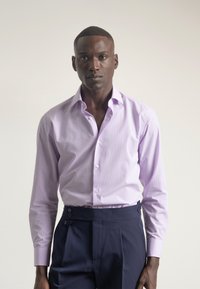 Camisa de vestir de manga larga color púrpura claro con cuello abotonado y un sutil patrón de rayas. Combinada con pantalones oscuros de talle alto de color azul marino con botones laterales.