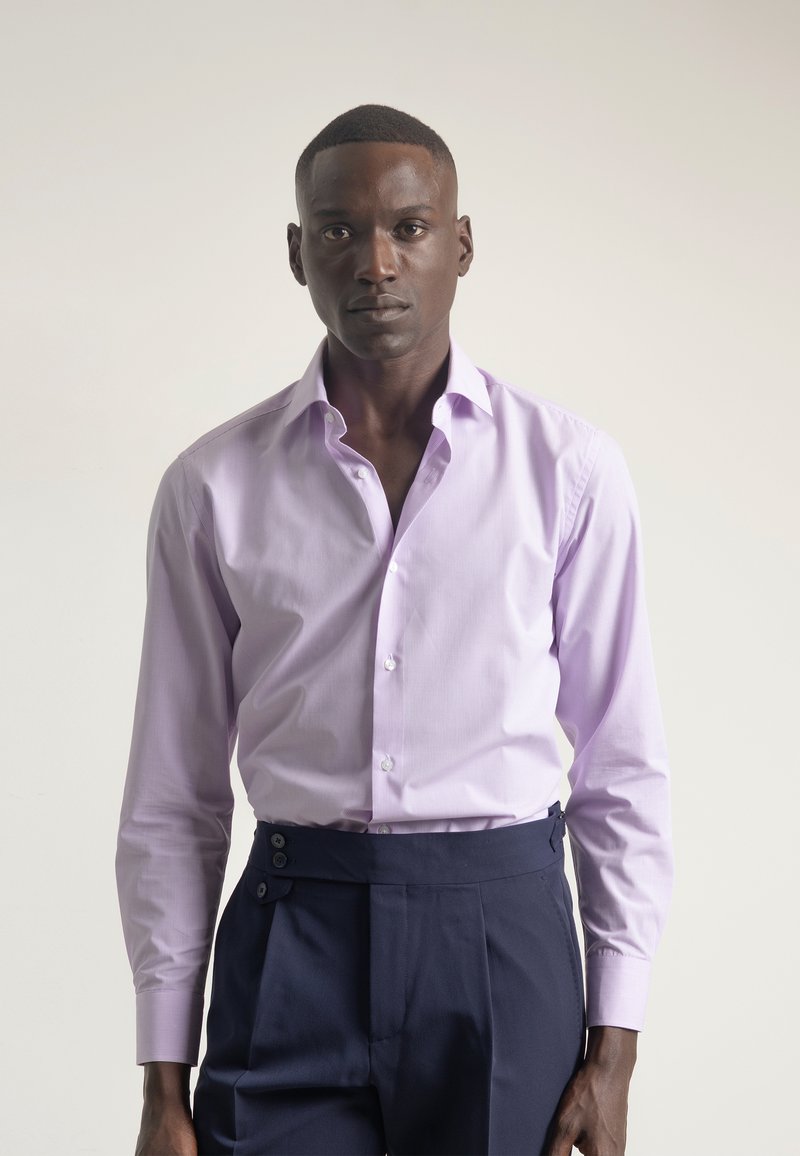 Camisa de vestir de manga larga color púrpura claro con cuello abotonado y un sutil patrón de rayas. Combinada con pantalones oscuros de talle alto de color azul marino con botones laterales.