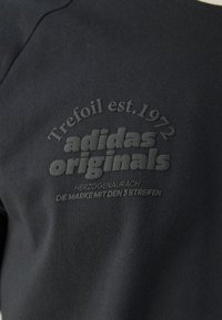 Černá látka s vystouplým černým textem „Trefoil est. 1972 adidas originals Herzogenaurach Die Marke mit den 3 pruhy.“