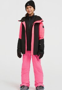 Roze en zwarte geïsoleerde skijas met een rits aan de voorkant en een capuchon, gecombineerd met roze skibroeken en zwarte handschoenen. Sneeuwschoenen zichtbaar onderaan.