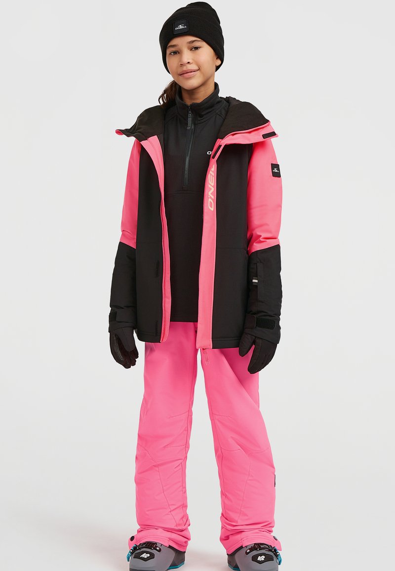 Roze en zwarte geïsoleerde skijas met een rits aan de voorkant en een capuchon, gecombineerd met roze skibroeken en zwarte handschoenen. Sneeuwschoenen zichtbaar onderaan.