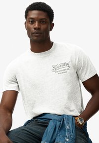 Camiseta de algodón gris con mangas cortas y cuello redondo. Presenta un logotipo de "Superdry" impreso en gris oscuro en el pecho.
