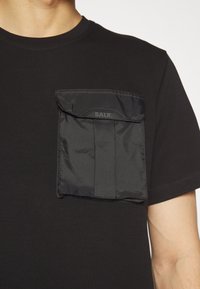 Svart t-shirt med en texturerad, svart tygficka som har en klaff och "BALR."-märke. Den har en klassisk rund halsdesign.