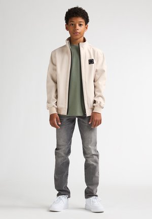 Beige bomberjack met zwarte rits en logo, gedragen over een groen T-shirt. Gecombineerd met grijze spijkerbroek en witte sneakers. Overal een gladde textuur.