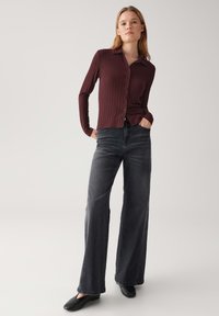 Maglietta a maniche lunghe a coste color amaranto con bottoni sul davanti, abbinata a jeans a gamba larga grigio scuro a vita alta e scarpe nere senza lacci.