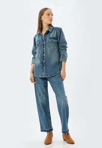 Camisa de denim y jeans de pierna ancha en un lavado azul medio. La camisa tiene cierre de botones, bolsillos en el pecho y un corte relajado. Los jeans tienen un dobladillo deshilachado.