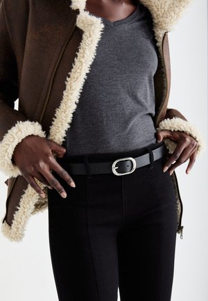 Braune mit Schaffell gefütterte Jacke, graues V-Ausschnitt-Shirt, schwarzer Gürtel mit ovaler silberner Schnalle, schwarze enge Jeans, strukturierte Materialien und geschichtetes Design.
