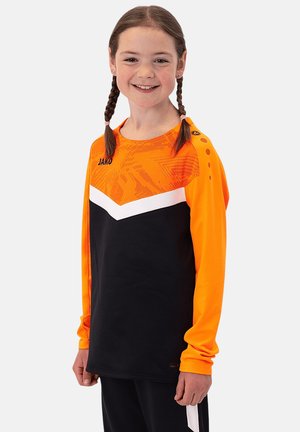 Ragazza sorridente con capelli intrecciati che indossa una maglia sportiva a maniche lunghe arancione e nera con un motivo a chevron bianco.