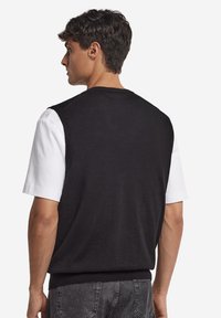 Svart stickad väst över en vit kortärmad skjorta, med rund halsringning och ribbad fåll, som har ett texturerat stickmönster.