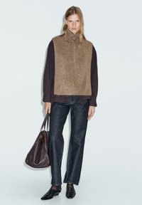 Gilet in pile marrone con colletto alto sopra una maglietta scura a maniche lunghe. Jeans in denim scuro con vestibilità ampia e scarpe nere a punta. Borsa a mano marrone.
