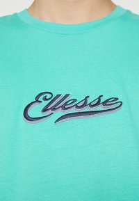 T-shirt en coton turquoise avec logo « Ellesse » brodé en noir et rose sur le devant. Encolure ronde avec tissu texturé.
