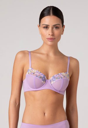Donna che indossa un reggiseno lavanda con ricamo floreale sulle coppe superiori su uno sfondo chiaro uniforme.