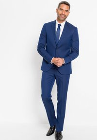 Abito blu navy con una texture liscia, chiusura a un bottone e rever a punta. Abbinato a una camicia bianca e una cravatta scura, completato con scarpe nere.