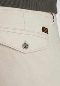 Los pantalones de algodón beige cuentan con un bolsillo trasero con un botón metálico y una etiqueta de tela negra con texto dorado, creando un diseño texturizado.