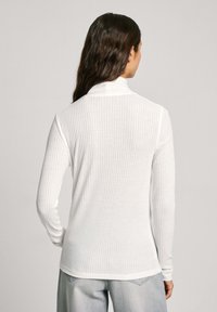 Top à manches longues blanc à côtes, avec un col haut et un tissu texturé. L'ourlet est arrondi et il est associé à un jean en denim gris clair.