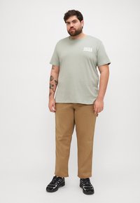Lys grønn t-skjorte med "JACK & JONES" logo i hvitt, kombinert med beige bukser og svarte joggesko med aksentfarger. Casual fit.
