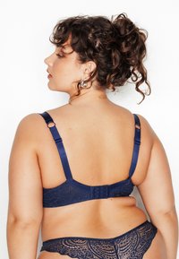 Reggiseno in pizzo blu navy con spalline in raso, regolabili, e un pannello decorativo sul retro. Il design presenta motivi in pizzo intricati.