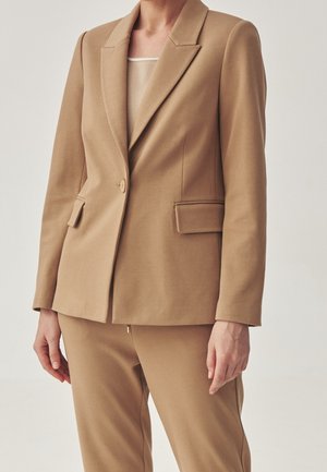 Femme portant un blazer camel coupé sur mesure avec un bouton unique et un pantalon assorti sur un fond clair uni.