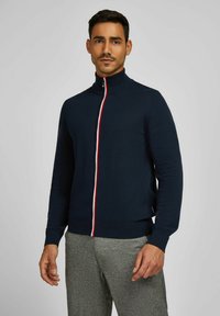 Donkerblauwe gebreide zip-up trui met een hoge kraag, voorzien van een rode en witte streep langs de rits. Aansluitende geribbelde manchetten en onderrand.