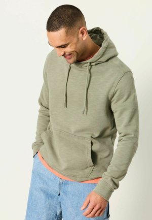 Man smiling downward wearing olive green hoodie with front pocket and light blue jeans against plain background.