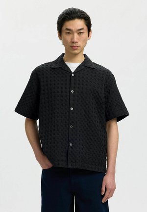 Mężczyzna w czarnej teksturowanej koszuli z krótkim rękawem zapinanej na guziki, na białej koszulce, stojący z jedną ręką w kieszeni na jednolitym tle.