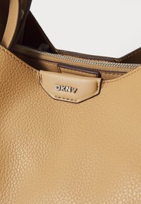 Sac en cuir tan avec une texture granuleuse, arborant une étiquette de marque DKNY. Il possède un compartiment zippé et des accents marron lisses.