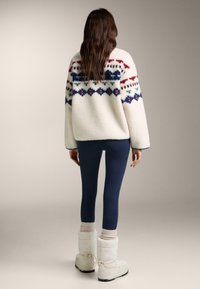 Vit fleecetröja med flerfärgade geometriska mönster, kombinerad med marinblå leggings och vita fluffiga stövlar. Enkel, mysig look.