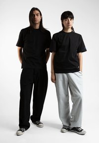 To personer står side om side foran en hvid baggrund. Den ene har en sort t-shirt og bukser på, mens den anden har en sort t-shirt og grå sweatpants.