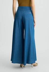 Weite Denim-Hose in einem mittleren Blauton. Mit hoher Taille, zwei Gesäßtaschen und einer glatten Textur. Mit einem einfachen Stickdetail gefertigt.