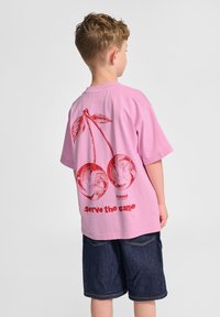 En rosa bomullströja med en röd körsbärsbild tillsammans med blad och texten "serve the game." Barnets rygg är synlig och de har på sig mörka denimshorts.