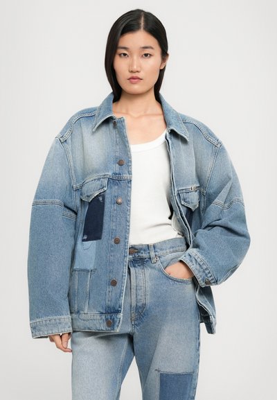 Mladá žena v oversized světle modré patchwork džínové bundě, odpovídajících džínách s vysokým pasem a bílé tílko, stojící na pozadí bez vzoru.