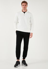 Hanorac alb cu fermoar, pantaloni de jogger negri și adidași negri. Material neted, croi standard, cu manșete și tiv zimțat. Design simplu.