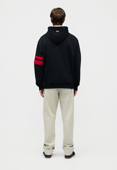 Mann som står med ryggen til, iført en svart hettegenser med røde striper på venstre arm og lyse beige bukser med svarte sko.