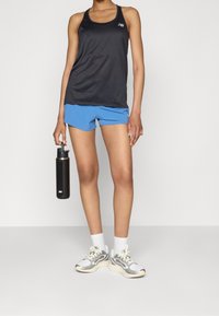 Black sports tank top with racerback design, paired with blue shorts. Holding a black water bottle, wearing light gray and cream sneakers.