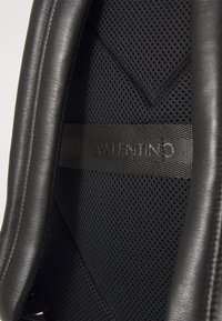 Binnenkant van een zwart leren en mesh rugzak met een gladde textuur, gevoerde delen, en een gebrandmerkte zwarte schouderband met "Valentino" in reliëf.