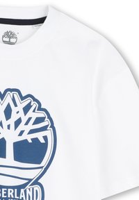 Bijela pamučna majica s plavim okruglim logotipom s dizajnom drveta i natpisom "Timberland" ispod. Kratke rukave, okrugli izrez.