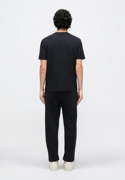 Čierne krátke tričko s textúrou a krátkymi rukávmi, v páre s čiernymi teplákmi, s rovnou lemou a voľným strihom. Model stojí na bielom pozadí.