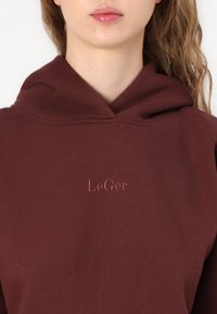 Burgundská mikina s mäkkou textúrou, s kapucňou na šnúrku a vyšitým logom "LeGer" v svetlejšom odtieni nad oblasťou hrude.