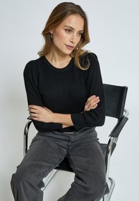 Črna pletena pulover s okroglo vratno linijo in naborki na rokavih, v kombinaciji s sivimi hlačami širokega kroja, sedeča na krom stol. Preprosti detajli teksture.