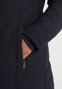 Marinblå puffjacka med en sidoficka med dragkedja, slät tygkvalitet och elastiska ärmmuddar för värme och komfort.