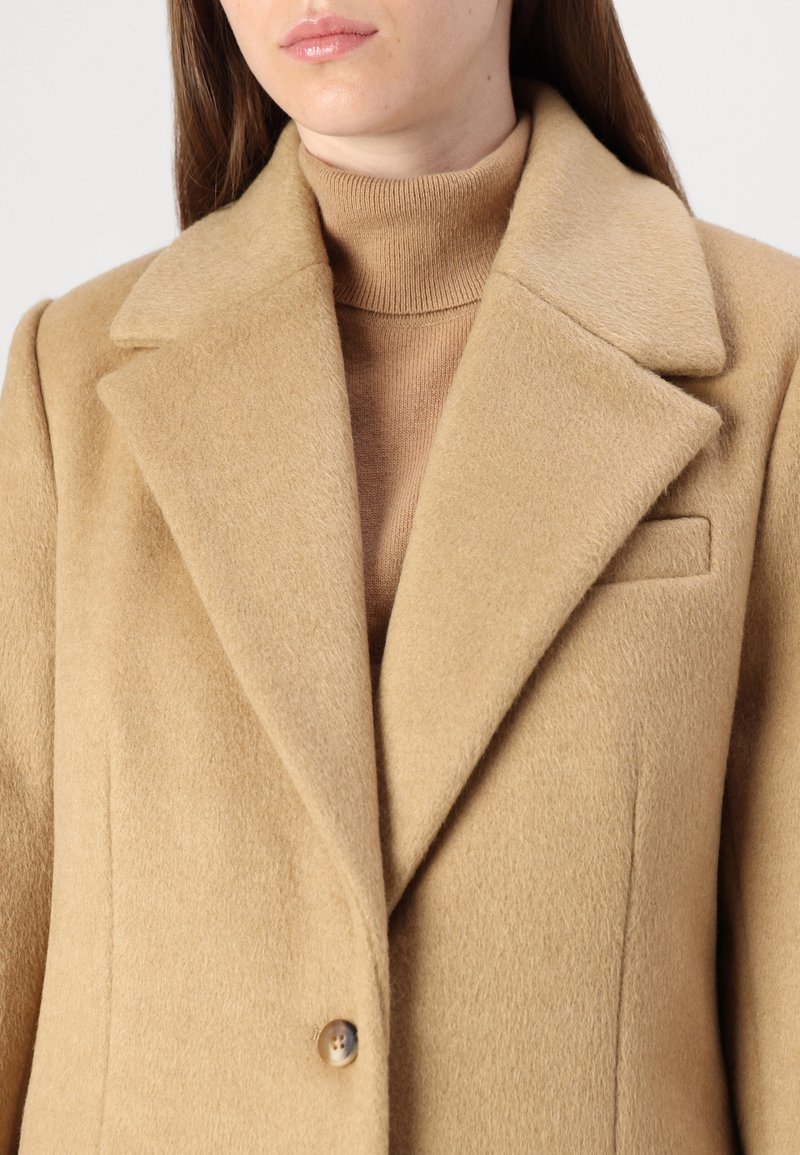 Płaszcz z mieszanki wełny w kolorze camel z klapami, z jednolitą zapięciem na guzik i gładką teksturą. Noszony na beżowym swetrze z golfem.