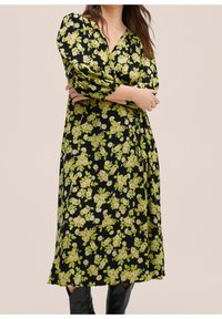 Robe noire à motifs floraux verts et crème, décolleté en V, manches 3/4 et ceinture à la taille, fabriquée dans un tissu léger et texturé.
