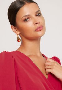 Kobieta z gładkimi, ciemnymi włosami spiętymi z tyłu, ubrana w czerwoną sukienkę z dekoltem w serek i złotymi, zakrzywionymi kolczykami w kształcie koła, trzyma materiał przy swoim ramieniu.